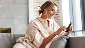 młoda kobieta siedzi uśmiechnięta w mieszkaniu na kanapie i korzysta ze smartfona
