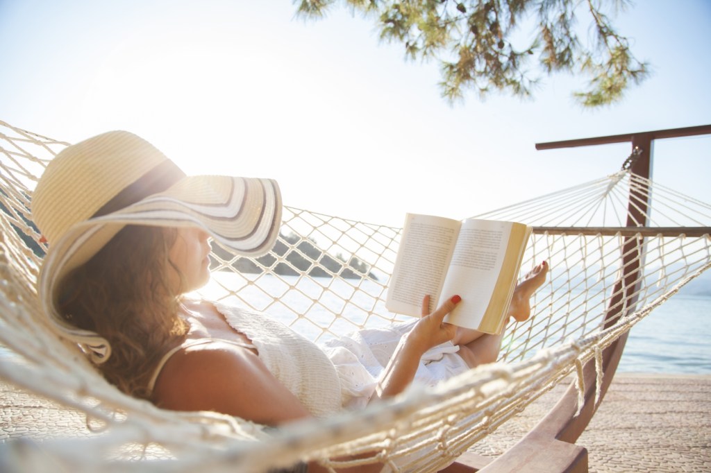 Kobieta w słomkowym kapeluszu leży w hamaku w pobliżu plaży w słoneczny letni dzień i czyta książkę